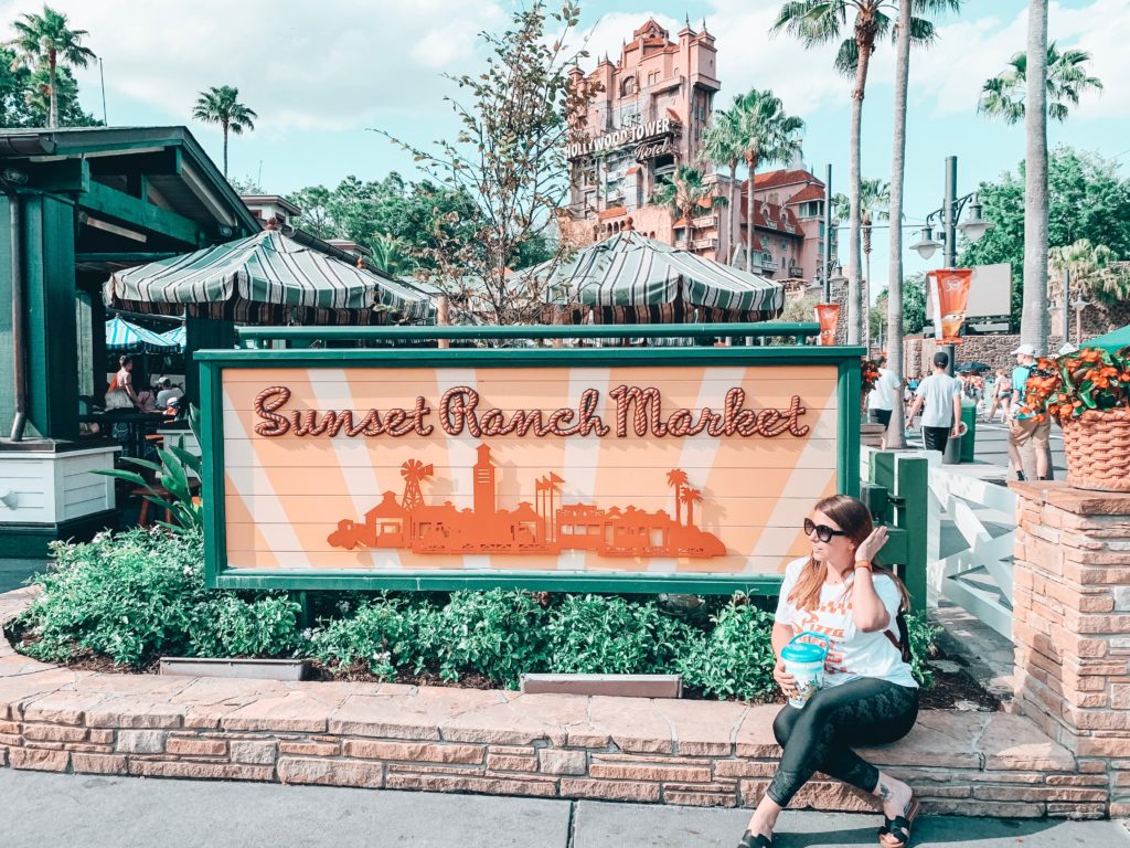 girl sitting in front of Sunset Ranch Market sign with the Hollywood Tower of Terror in the background at Disney's Hollywood Studios.